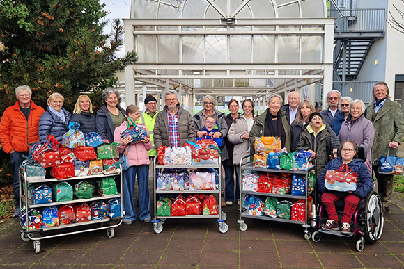 Gruppenfoto der Frauen und Männer der drei beteiligten Vereine gemeinsam mit der CHS-Schulleitung sowie Schülerinnen und Schülern hinter drei mit Weihnachtspaketen gefüllten Rollwägen